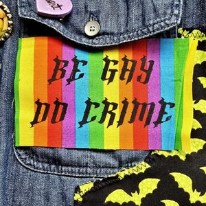 May include: A denim jacket with a rainbow patch that reads "BE GAY DO CRIME" in black gothic lettering. The jacket also features a pin with a gold border and a purple heart-shaped pin.