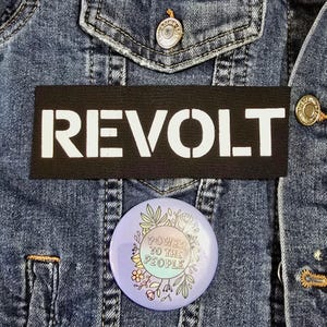 Peut inclure: Veste en jean bleue avec un patch rectangulaire noir portant l'inscription blanche "REVOLT". En dessous, un badge rond affiche "POWER TO THE PEOPLE" entouré de motifs floraux.