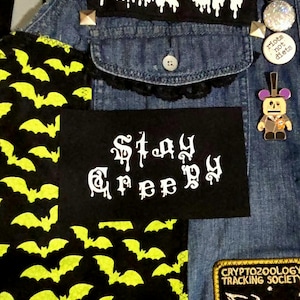 May include: Close-up of a denim jacket with patches and pins. A black patch with the words "Stay Creepy" in white gothic lettering is visible. Other patches feature bats and a Cryptozoology Tracking Society logo. Decorative pins and studs are also present.