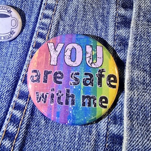 Puede incluir: Un botón de colores arcoíris con el texto "You are safe with me" en letras negras.
