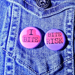 May include: Two round, colorful buttons with text on a blue denim jacket. One button is pink with the words "I BITE" in black and red. The other button is purple with the words "BITE RISK" in black and red.