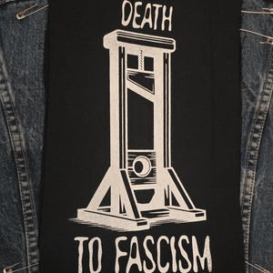 May include: Black patch with a white guillotine graphic and the text "DEATH TO FASCISM." The patch is rectangular and is attached to a denim jacket with safety pins. The graphic is a bold, stark design.