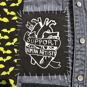 May include: A denim shirt with a black patch featuring a white heart design and the words "SUPPORT HUMAN ARTISTS." The patch is stitched onto the shirt with white thread. A black fabric with yellow bats is visible on the left side.