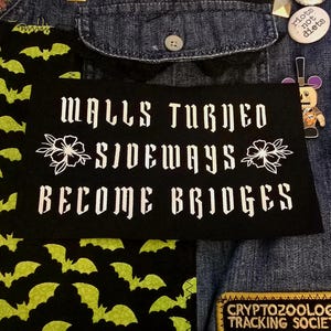 以下が含まれることがあります： 「WALLS TURNED SIDEWAYS BECOME BRIDGES」と白い文字と花の模様が描かれた黒いパッチ。他のパッチやピンが付いたデニムジャケットに配置されており、コウモリ柄の生地も含まれています。