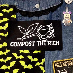 Puede incluir: Primer plano de una chaqueta vaquera con parches y alfileres. Se ve un parche negro con el texto "COMPOST THE RICH" y un diseño de rosa blanca. Otros parches incluyen un patrón de murciélagos y un pin de un robot.