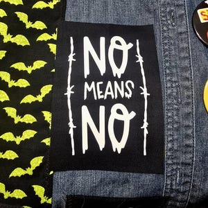 May include: Black patch with white text that reads "NO MEANS NO" surrounded by barbed wire. The patch is sewn onto a denim jacket.