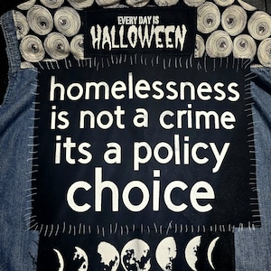 May include: A denim vest with patches. The top patch reads "EVERY DAY IS HALLOWEEN" in white text on a black background. The main patch says "homelessness is not a crime its a policy choice" in white text on a black background.