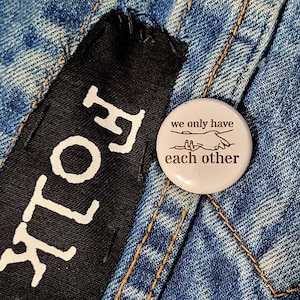 May include: A white button with a brown outline and text that reads "we only have each other". The button features a line drawing of two hands reaching towards each other.