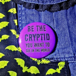 May include: A purple button with the text "BE THE CRYPTID YOU WANT TO SEE IN THE WORLD" in black dripping font. The button is set against a denim background with a black lace trim and a fabric with a bat pattern.