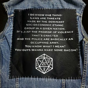 May include: A blue denim jacket with a black patch stitched on the back. The patch displays white text: "I DO KNOW ONE THING: LAWS ARE THREATS...YOU GUYS WANNA MAKE SOME BACON?" A white D20 die graphic is below the text.