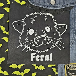May include: A black patch with a white cartoon cat face and the word "Feral" in white. The patch is surrounded by fabric with a bat pattern and denim fabric.