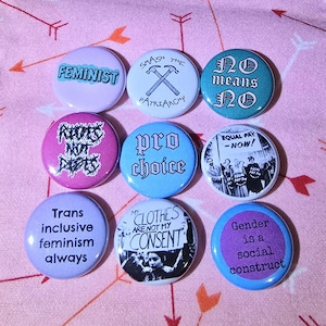 May include: A collection of nine circular feminist badges in pastel colours. The badges feature slogans such as "Feminist", "Smash the Patriarchy", "No means No", and "Pro Choice". The text "Gender is a social construct" is also visible.