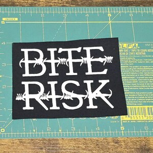May include: A black rectangular patch with the words "BITE RISK" in white, styled with barbed wire. The patch is on a green cutting mat with centimetre measurements.