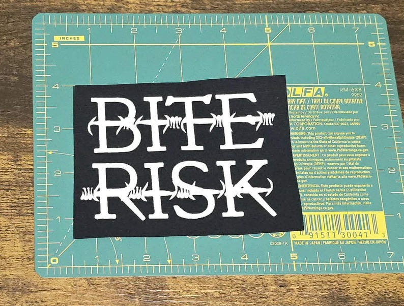 May include: A black rectangular patch with the words "BITE RISK" in white, styled with barbed wire. The patch is on a green cutting mat with centimetre measurements.