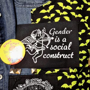 May include: A black patch with white embroidery depicting a cherub with an arrow and the text "Gender is a social construct". The patch is sewn onto a denim jacket with a green and black bat print fabric.