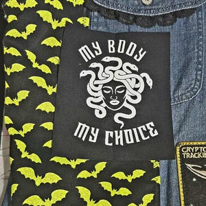 May include: A black patch with the text "MY BODY MY CHOICE" and a white Medusa illustration. The patch is on a denim jacket with a black bat pattern fabric. The bats are yellow.