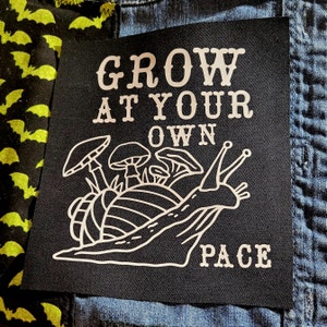 May include: Black patch with white text and a snail illustration. The text reads "GROW AT YOUR OWN PACE". The snail is surrounded by mushrooms.