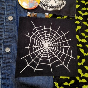 May include: A black fabric patch with a white spiderweb design. The patch is sewn onto a denim jacket with other patches.