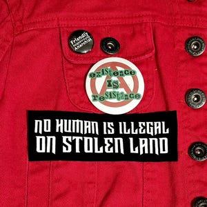 May include: A close-up of a red denim jacket adorned with patches and buttons. The jacket displays a black patch with white text: "NO HUMAN IS ILLEGAL ON STOLEN LAND." There's also a pin with the text "Existence Is Resistance" and two buttons.