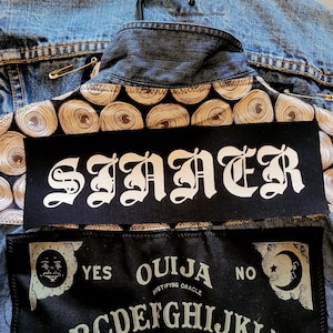 May include: A denim jacket with a black patch that reads "SINNER" in white gothic lettering. The jacket has a decorative pattern of eye-like shapes and a Ouija board design.