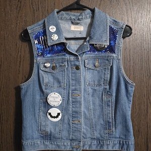May include: A blue denim vest with a galaxy print fabric on the collar and back. The vest has multiple pins and buttons, including a bat, a pin that says "Everyday is Halloween", and a pin that says "Cream & Teal".
