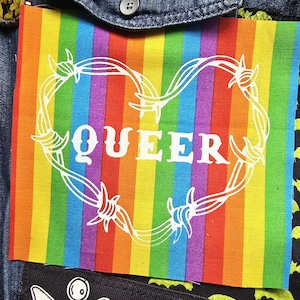 Peut inclure: Un écusson à rayures arc-en-ciel avec le mot "QUEER" en blanc, entouré d'un motif de cœur en fil de fer barbelé blanc. L'écusson est cousu sur une veste en jean, avec un tissu noir avec des silhouettes de chauves-souris vertes visibles à droite.