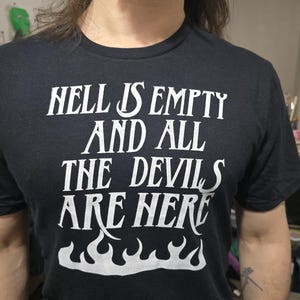 Puede incluir: Camiseta negra con el texto blanco "HELL IS EMPTY AND ALL THE DEVILS ARE HERE" sobre un diseño de llamas. La camiseta es de cuello redondo y parece estar hecha de un material suave.