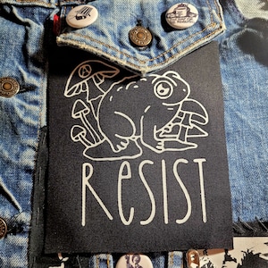 May include: Black patch with a white outline of a frog sitting on a mushroom with the word "RESIST" in white letters below.