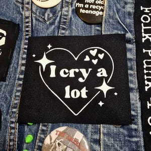 May include: Black iron-on patch with a white heart that says "I cry a lot" with stars and small hearts around it.