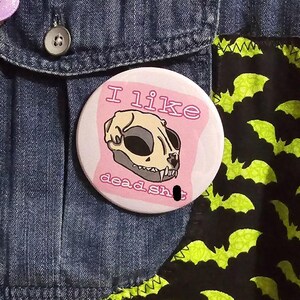 May include: A round button with the text "I like dead shit" and a cat skull illustration on a pink background. The button is on a denim jacket with a black fabric patch featuring green bat silhouettes.
