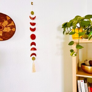 May include: A red and gold moon phase wall hanging with a tassel. The hanging is made of ceramic and features a series of crescent moons in different phases.