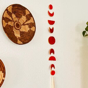 May include: A red wooden moon phase wall hanging with a white tassel. The moon phases are in various stages of fullness, from a full moon to a crescent moon.