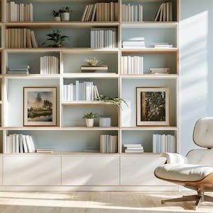 Könnte beinhalten: Ein großes, deckenhohes weißes Bücherregal mit hellen Holzakzenten, gefüllt mit Büchern, kleinen Topfpflanzen und zwei gerahmten Landschaftsbildern. Ein weißer Ledersessel mit Holzrahmen steht rechts neben dem Regal.