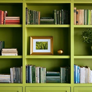 May include: A large, olive green built-in bookshelf featuring three vertical sections with multiple shelves. The shelves are filled with various books organized by color, a gold-framed picture, a small gold sphere, and a potted green plant.