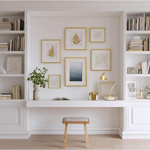 May include: A white fitted bookcase with a desk and a small wooden stool. The desk features framed art, a lamp, and a vase with greenery. Bookshelves are filled with books and decorative items. The wall art has gold frames.