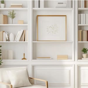 Puede incluir: Estanterías blancas empotradas con molduras blancas y detalles dorados. Las estanterías están llenas de libros, objetos decorativos y plantas. Un grabado enmarcado con un marco dorado cuelga de la estantería del medio.
