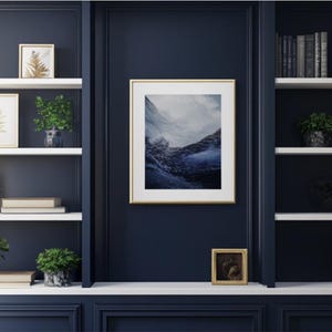 May include: A white bookshelf with three shelves, each filled with books, plants, and decorative items. The bookshelf is against a dark blue wall. A framed photograph of a mountain landscape hangs on the wall above the bookshelf.