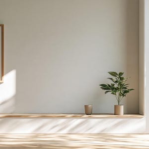 以下が含まれることがあります： 緑の植物プリントが施された額入りのウォールアートが、長い木製の棚の上に掛けられています。棚の上には小さなベージュのカップと鉢植えの緑の植物が置かれています。大きな窓から差し込む日光が床に影を落としています。