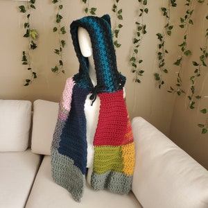 Può includere: Una sciarpa con cappuccio all'uncinetto colorata con un design a patchwork in tonalità di blu, verde, rosso, rosa e grigio. La sciarpa è esposta su un divano bianco.