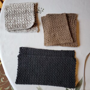 Puede incluir: Tres artículos de ganchillo hechos a mano en colores neutros se exhiben sobre un mantel blanco. Los artículos incluyen un cuadrado plegado moteado gris claro y blanco, un cuadrado plegado marrón y una pieza rectangular gris oscuro. Los artículos están hechos de hilo texturizado.