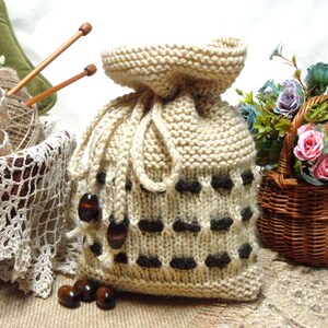Könnte beinhalten: Eine beige gestrickte Kordeltasche mit braunen Akzenten und einem karierten Muster. Die Tasche liegt auf einer braunen Oberfläche mit Holzperlen und einem Blumenkorb.