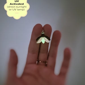 以下が含まれることがあります： 暗い色のホタル型のペンダントで、緑色の光るアクセントが付いています。ペンダントはニュートラルな背景に表示されています。「UV Activated」というテキストが黄色のバブルに見えます。ペンダントは直射日光またはUV光の下で光るように設計されています。