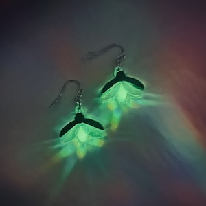 Op de afbeelding: Een paar zilveren oorhangers met een groen oplichtend bloemetjesontwerp.