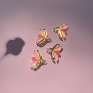 May include: Four gold and pink butterfly-shaped hair clips on a pink background.