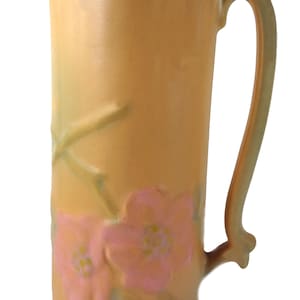 Puede incluir: Una jarra de cerámica vintage con un esmalte amarillo pálido y un diseño floral rosa. La jarra tiene una boca ancha y un cuerpo alto y cilíndrico con una base redondeada.