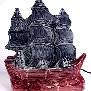 Puede incluir: Una lámpara de cerámica en forma de barco con un barco gris oscuro y una base roja. El barco tiene tres velas y está decorado con cuerdas y aparejos. La base está decorada con olas.