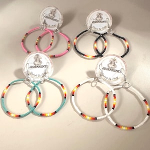 May include: Four pairs of beaded hoop earrings in pink, black, turquoise, and white. Each pair features a circular design with a beaded pattern in various colors, including red, yellow, and black. The earrings are attached to circular tags with the text "ADORNMENTS".