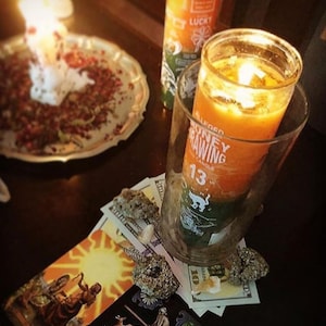 May include: A lit orange and green candle in a glass, labeled "Alleged Money Drawing 13", sits on a table with money, crystals, and a tarot card. Another candle labeled "Lucky" is in the background. A plate with red and white items is also visible.