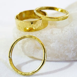 May include: Three gold rings, two with a hammered texture and one smooth, stacked on a white rock.