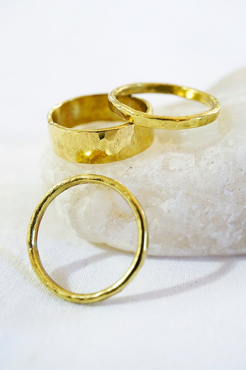May include: Three gold rings, two with a hammered texture and one smooth, stacked on a white rock.